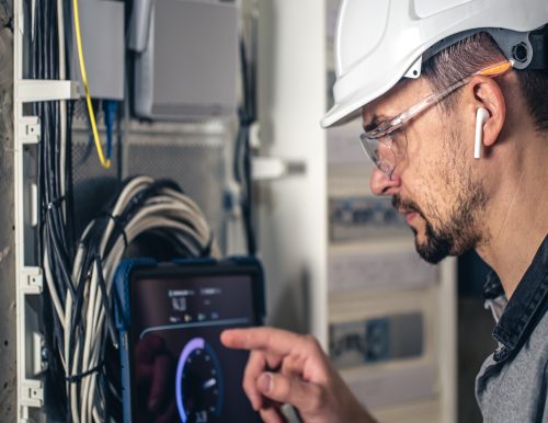 Man, an electrical technician working in a switchboard with fuses. Installation and connection of electrical equipment. Professional uses a tablet.